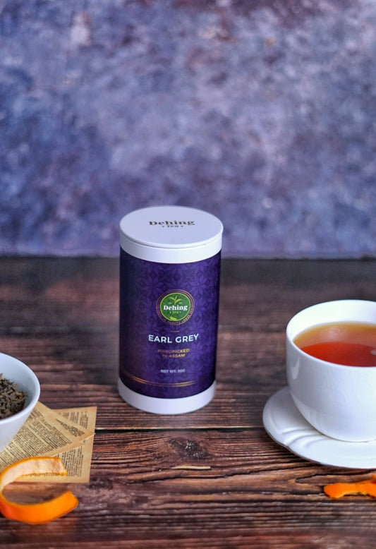 A wooden surface with a freshly brewed cup of tea alongside a gift caddy cannister of Dehing Tea's Earl Grey loose leaf tea and bergamot rind