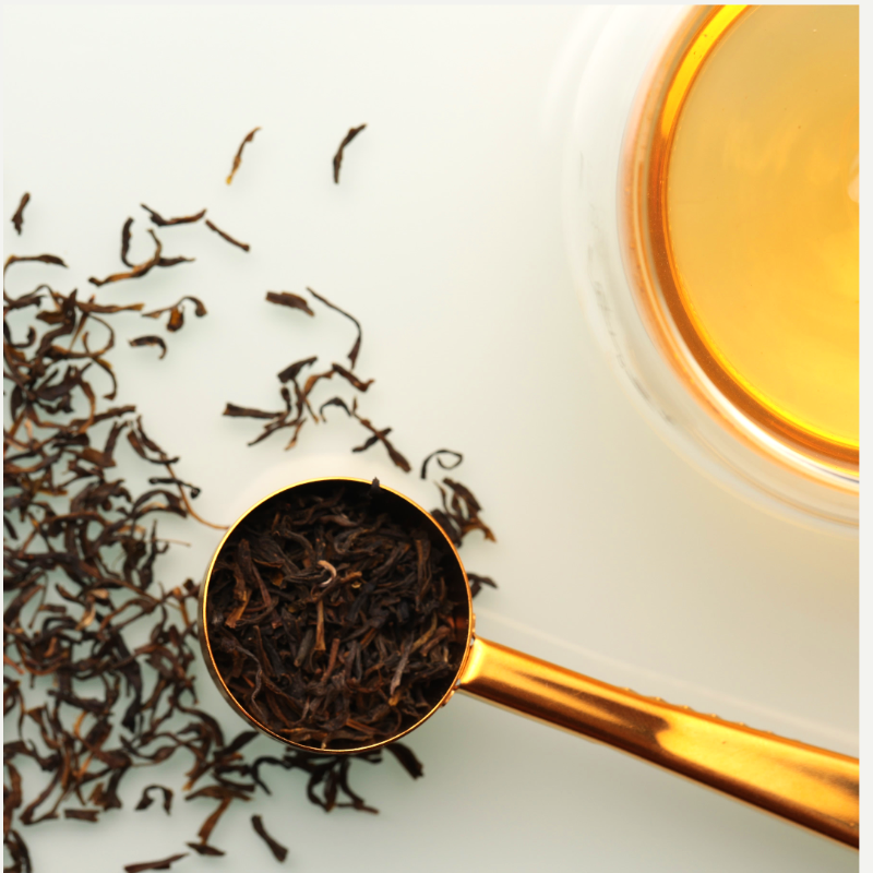 Scoop of Assam loose leaf black tea with scattered tea leaves on the surface alongside a brewed cup of golden tea