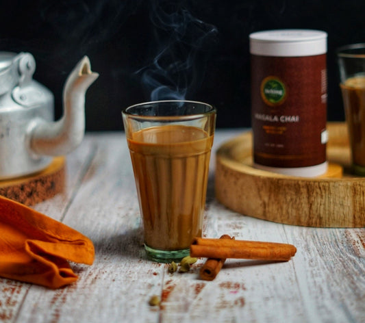 Freshly brewed masala chai tea in a glass with cinnamon sticks on the table next to it and a pot of tea and Dehing gift caddy of Masala Chai loose leaf tea in the background