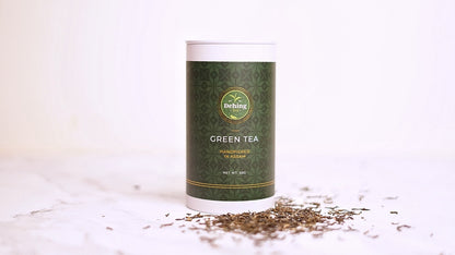 Dehing tea caddy labelled 'Green Tea' with scattered tea leaves on a light background
