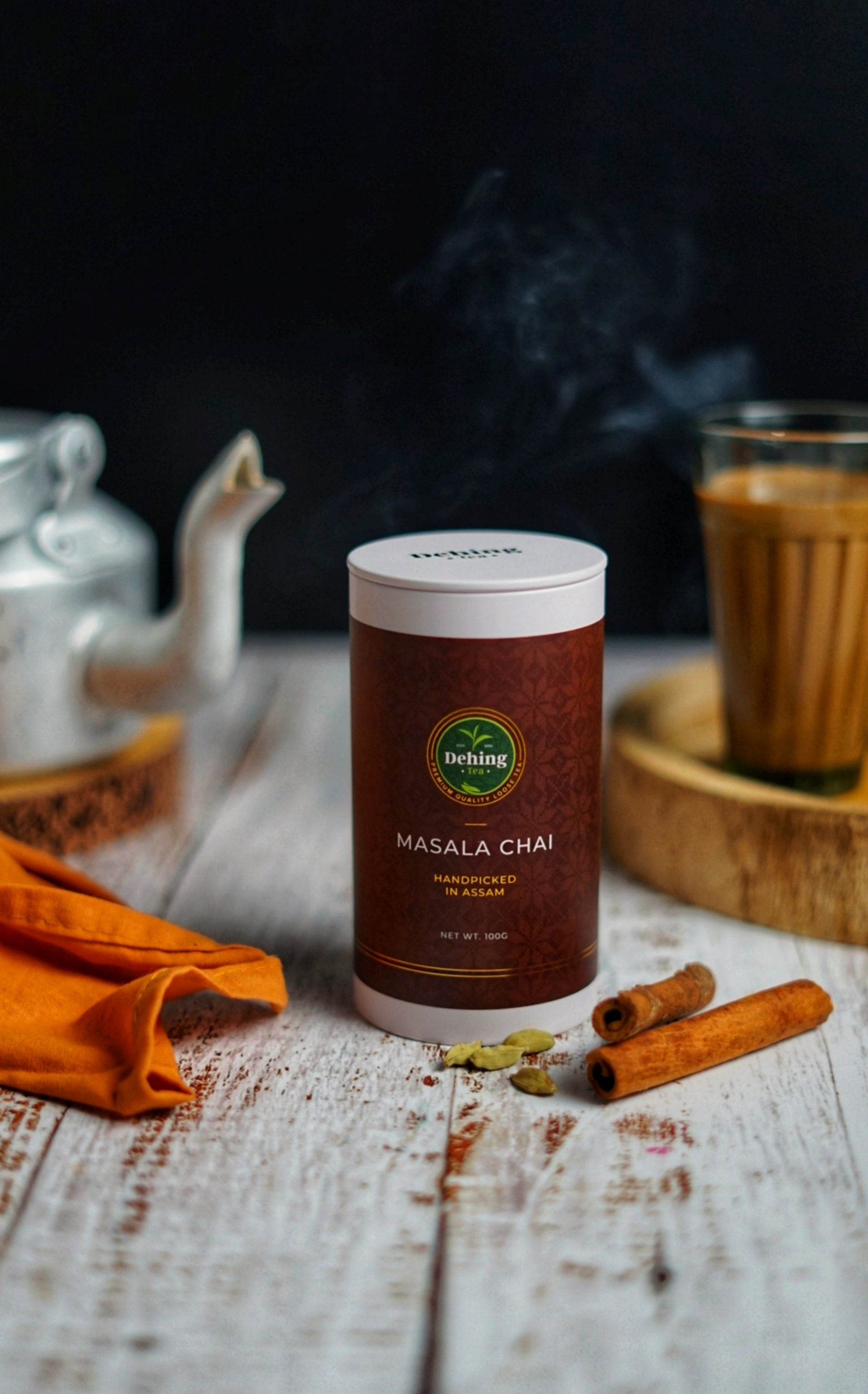 Canister gift caddy of Masala Chai on a wooden surface with cinnamon sticks and cardamom spices and a glass of tea  with tea pot in the background.