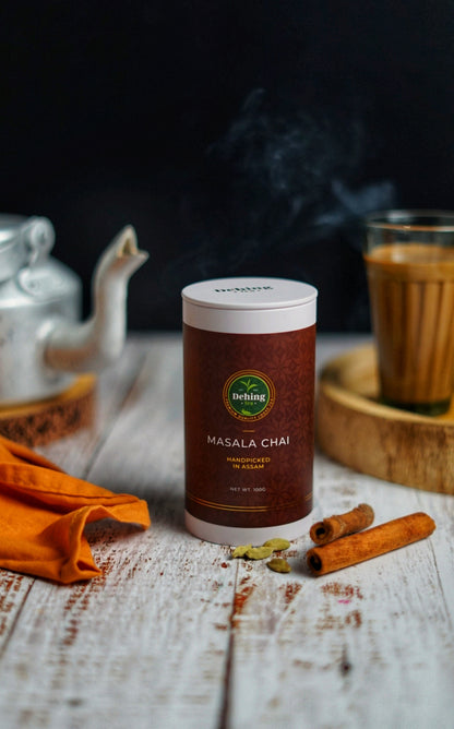 Canister gift caddy of Masala Chai on a wooden surface with cinnamon sticks and cardamom spices and a glass of tea  with tea pot in the background.