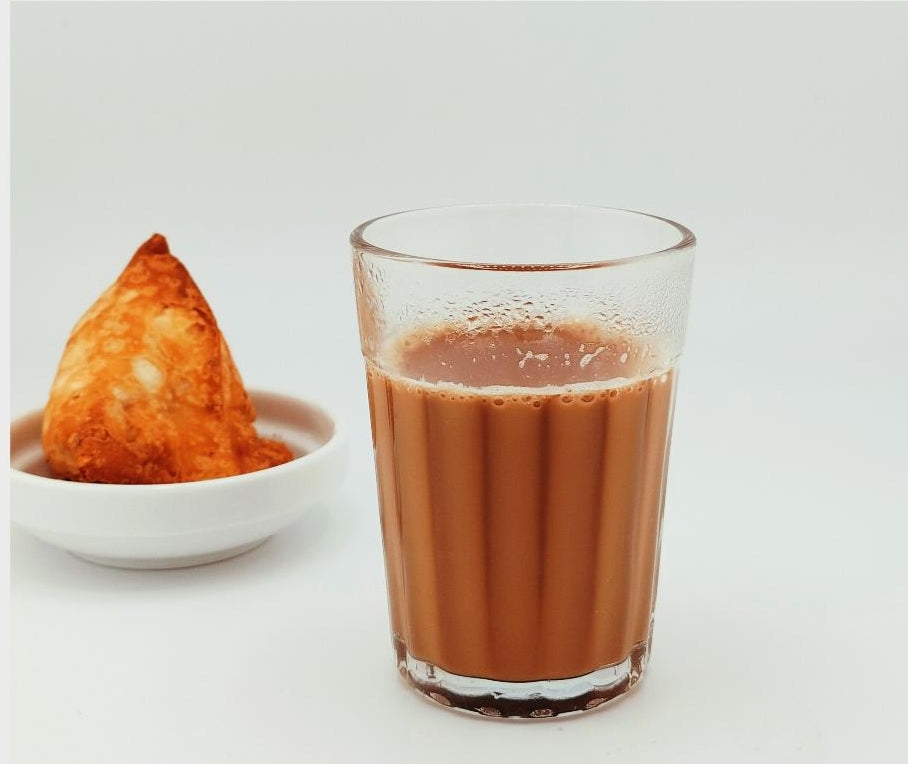Freshly brewed and ready to drink masala chai tea in a glass with a cooked samosa in the background